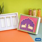 A white book-shaped hamper box featuring purple and pink Pichwai art with three glass-like jars and gold lids.